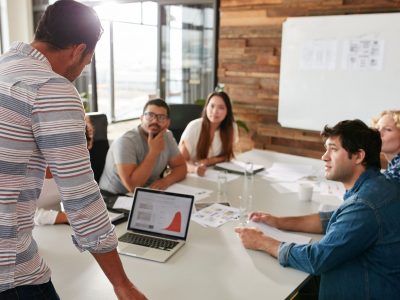 Young man giving business presentation on laptop to colleagues sitting around table in conference room.